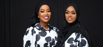 Two women wearing black hijabs and matching white dresses with bold black floral prints, smiling