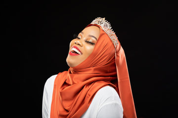 Woman in orange satin hijab and jeweled crown, smiling, against a black background