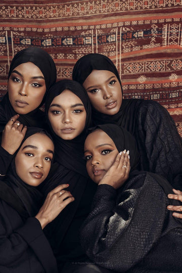 Five women wearing black hijabs and elegant modest clothing posing together in front of a patterned rug background.
