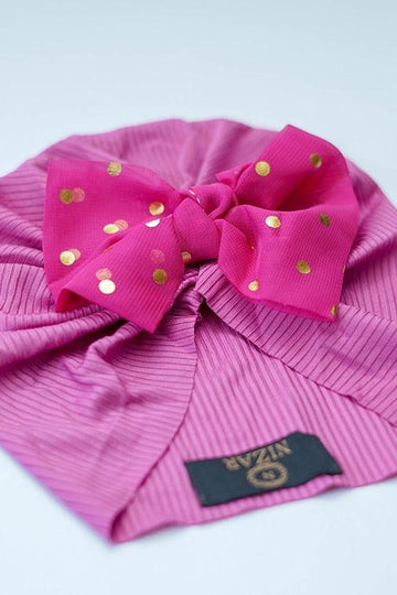 Pink ribbed baby turban with large bow and gold polka dots on a light background