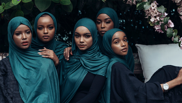 Five women in teal hijabs and modest dresses, seated together outdoors under greenery