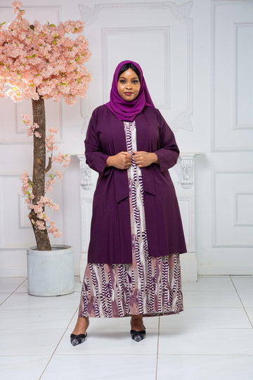 Woman in a purple hijab and abaya dress stands by a pink blossom tree in a bright room.